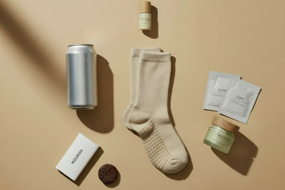 Create me a birds eye view aesethic image with beige back ground and shadwoing, it should include pilates socks, silver can, neutral wellness bar in packet, mini skincare, eye face mask,  little wellness treat